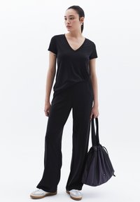 Top corto nero con scollatura a V abbinato a pantaloni neri a gamba larga. Il modello tiene una borsa a coulisse viola. Le scarpe sono bianche con dettagli grigi.