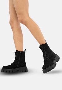 Botas negras de ante hasta el tobillo con una suela gruesa y estriada, parte delantera con cordones y cremallera lateral para facilitar su uso. Cuenta con una lengüeta en la parte posterior.