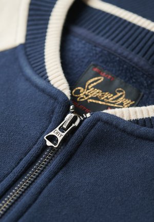 Granatowa bluza zapinana na zamek błyskawiczny z żeberkowym kremowym kołnierzem; posiada srebrny zamek z wygrawerowanym napisem "DRY" oraz naszywkę z logo w kolorze złotym.