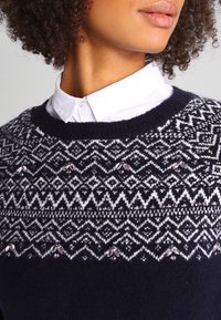 Marineblaue Strickpullover mit einem weißen geometrischen Muster, verziert mit kleinen Verzierungen entlang des Halsausschnitts und der Schultern.