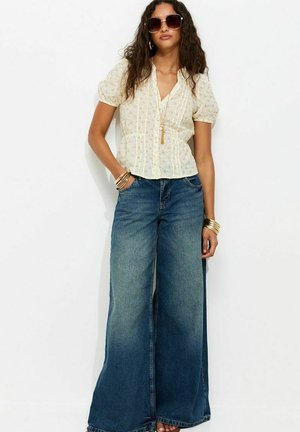 Kobieta nosząca duże okulary przeciwsłoneczne, kremową kwiecistą bluzkę z bufkami, szerokie niebieskie dżinsy i złote bransoletki na białym tle.