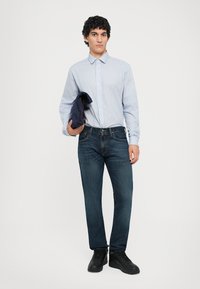 Light blue striped button-up shirt, dark denim jeans, black sneakers; model holds a navy jacket, standing against a white background.