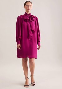Robe fuchsia avec de longues manches bouffantes et un grand nœud au niveau du col. Texture lisse, longueur au genou, associée à des talons noirs à fines brides.