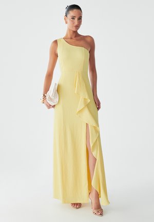 Model wearing a one-shoulder yellow dress with a side ruffle and slit, holding a small white clutch, against a plain background.