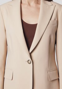 Beżowa marynarka o dopasowanym kroju, z pojedynczym zapięciem na guzik, klapami notch oraz dwiema przednimi kieszeniami. Gładka faktura i wyraźny kształt.