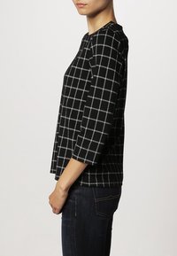 Žena v černém dlouhém tričku s bílým mřížkovým vzorem a tmavých džínách, stojící bokem na jednoduchém světlém pozadí.