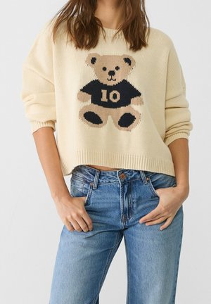 Kvinde iført cremefarvet strikket sweater med teddybjørn iført sort skjorte med nummer 10, kombineret med blå jeans, hænderne i lommerne.