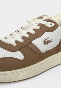 Baskets Lacoste blanches et marron avec lacets blancs, logo crocodile sur le côté, et semelle couleur crème sur un fond uni clair.