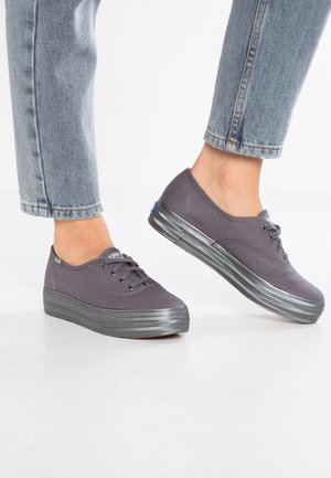 Personne portant des baskets compensées en toile grise et un jean bleu clair retroussé, debout sur un sol blanc.