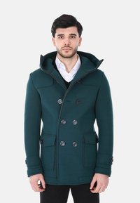 Uomo che indossa un cappotto doppiopetto verde scuro con cappuccio, camicia bianca e maglione nero, in piedi davanti a uno sfondo chiaro e uniforme.