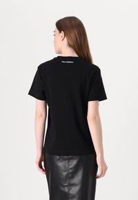 Černé tričko s krátkým rukávem a kulatým výstřihem, se znakem "KARL LAGERFELD" bílou barvou na zadní straně krku. Hladká textura látky.