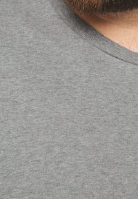 Gros plan sur le bas du visage d'un homme avec une barbe et une moustache brunes, portant un t-shirt gris à col rond.