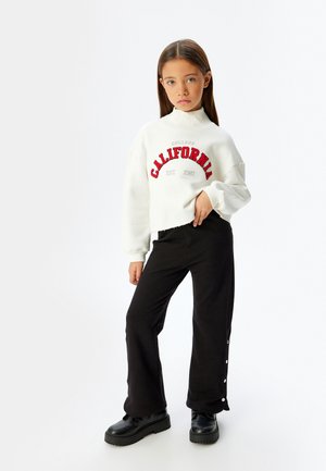 Sudadera blanca corta con letras rojas que dicen "CALIFORNIA", combinada con pantalones negros acampanados que presentan detalles de botones. Botas de plataforma negras.