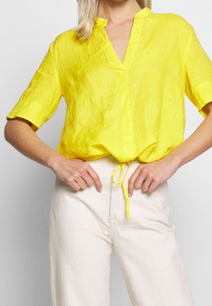 Kobieta ubrana w jasnoszosłtą bluzkę z krótkim rękawem, dekoltem w serek i wiązaniem w talii, zestawioną z wysokim stanem białymi spodniami.