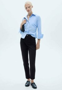 Svetlo modra srajca s kratkimi rokavi, združena z visokimi črnimi kavbojkami in črnimi loaferji. Črna torba se drži v desni roki.