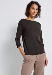 Mujer con una blusa negra de manga larga y escote barco, y pantalones beige con franjas laterales blancas, de pie contra un fondo claro y liso.