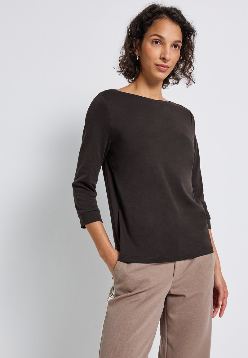 Mujer con una blusa negra de manga larga y escote barco, y pantalones beige con franjas laterales blancas, de pie contra un fondo claro y liso.