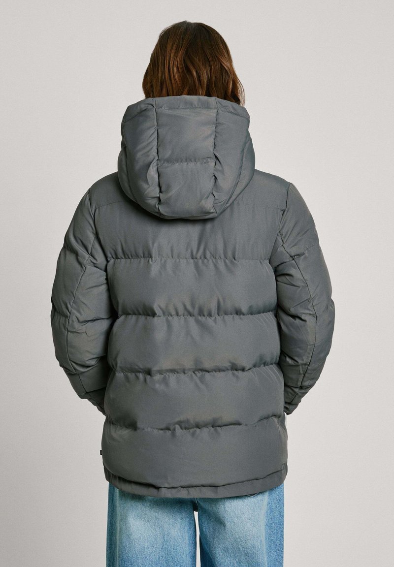 Giubbotto a vento grigio con cappuccio, caratterizzato da sezioni trapuntate e una superficie liscia, indossato sopra jeans azzurri chiari.