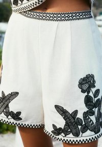 Bjele lanene kratke hlače s crnim cvjetnim vezom i obrubom, koje prikazuju vezeni pojas i detaljne biljne motive duž rubova.