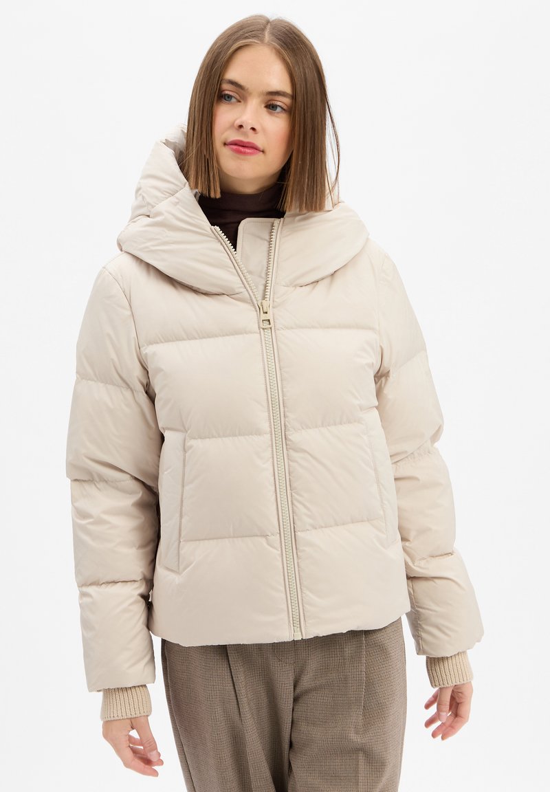 Woman with straight brown hair wearing a beige puffer jacket, brown turtleneck, and plaid pants, looking slightly to the side.