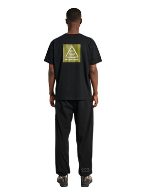 Person iført sort T-shirt med grønt firkantet logo med teksten "223 HALO OFF DUTY GEAR" og sorte bukser med teksten "223" nær anklen, stående med ryggen til.