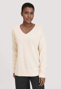 Cremefarbenes V-Ausschnitt-Pullover aus weichem Material mit langen Ärmeln sowie gerippten Bündchen und Saum; abgebildet an einem Model vor einem neutralen Hintergrund.