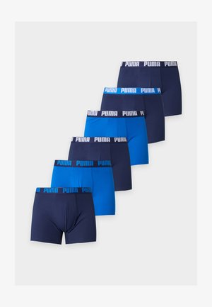 Atletyczny mężczyzna stoi boso, ubrany w ciemnoniebieskie bokserki Puma z niebieskim pasem, patrzy w lewo na jednolitym białym tle.