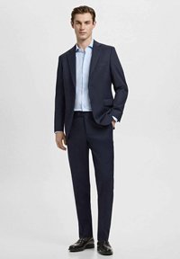 Mornarsko modro obleko s prisrčno prilegajočim krojem, enorazrezno srajco in ujemajočimi hlačami. Pod njo svetlomodra srajca in spolirani črni čevlji.