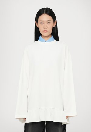 Witte oversized top met een gerimpelde zoom, met lange mouwen en een hoge kraag. Een lichtblauwe shirtkraag piept eronderuit.