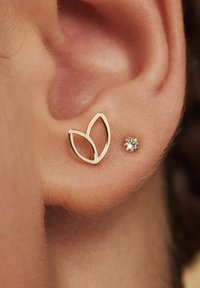 Goldene Metallohrringe mit blattförmigem Design und einem kleinen runden Kristallstein. Nahaufnahme eines Ohres, die Details und Texturen zeigend.