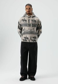 Beige capuchontrui met zwarte patronen van paarden en cactussen. Voorzien van trekkoorden en een losse pasvorm. Gecombineerd met een zwarte broek en robuuste schoenen.