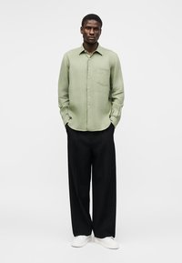 Άνδρας που φορά ανοιχτοπράσινο πουκάμισο με κουμπιά, μαύρο παντελόνι με φαρδιά πόδια και λευκά αθλητικά παπούτσια, στέκεται με τα χέρια στις τσέπες σε απλό φόντο.