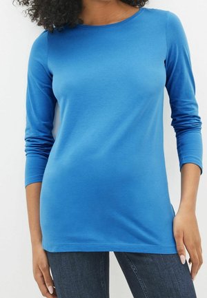 Femme portant un t-shirt bleu uni à manches longues et un jean foncé, debout, bras détendus le long du corps, sur un fond blanc.