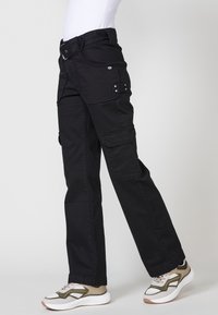Pantalones cargo negros hechos de tela duradera con múltiples bolsillos, detalles de botones y un ajuste relajado, combinados con zapatillas deportivas blancas y oliva.