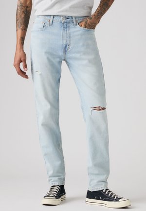 Homme portant un jean déchiré bleu clair et des baskets montantes noires, debout avec une main dans la poche et les bras tatoués visibles.