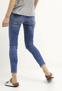Skinny jeans in blue denim, featuring a mid-rise waist and back pockets. Paired with black and silver sandals on a plain background.