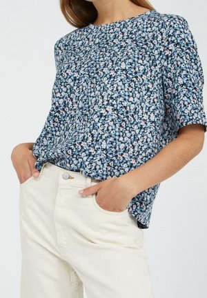 Femme portant un haut à manches courtes bleu à motifs floraux, rentré dans un pantalon taille haute blanc cassé, les mains dans les poches.