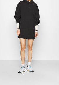 Schwarzes Kapuzensweatkleid mit einer Vordertasche, weiten Ärmeln und einem weißen Langarmshirt darunter. Kombiniert mit mehrfarbigen Sneakers.