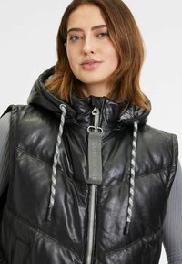 Schwarze Leder-Puffer-Weste mit einem Reißverschluss vorne, verstellbare Kapuze, grauen Kordeln und einer vorderen aufgesetzten Tasche. Seidige Textur und modernes Design.