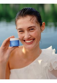 Blauwe cosmetica-pot met het label "LUMENE Nordic Hydration Moisturizer" vastgehouden door een persoon in een witte, gekruide top, met water op de achtergrond.