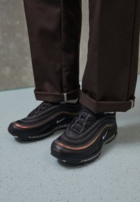 Person som bär svarta Nike Air Max-sneakers med kopparfärgad detalj, tillsammans med mörkbruna uppvikta byxor på ett fläckigt golv.