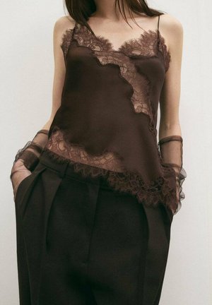 Camiseta de seda marrón con delicado borde de encaje en el escote y el dobladillo, de corte suelto y tirantes finos ajustables. Combinada con pantalones oscuros.