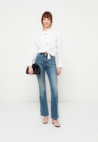 Camicia bianca con bottoni annodata in vita, jeans svasati blu, pochette nera e ballerine color rosa pallido; stile minimalista con un'estetica pulita.