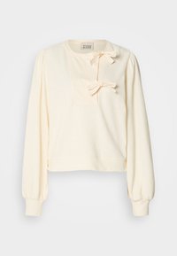 Sweatshirt couleur crème avec des manches longues bouffantes, présentant un patch texturé à l'avant et deux nœuds décoratifs au niveau du col.