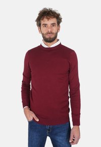 Maglione bordeaux a girocollo, texture lavorata a maglia, design aderente, indossato sopra una camicia bianca con colletto, abbinato a jeans di denim blu, mani nelle tasche.