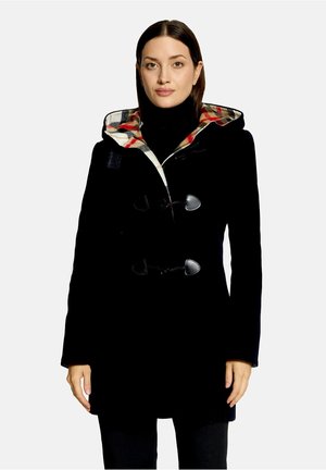Femme portant un manteau duffle-coat noir avec une doublure à carreaux et des fermetures à brandebourgs, debout devant un fond clair uni.