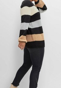 Persona che indossa un maglione a maglia a righe nero, beige e bianco, con jeans neri e stivaletti beige, che cammina su uno sfondo neutro.