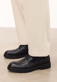 Must mustad nahked, ümarad varbad, tekstuuriga ülemine osa ja paeltega disain; koos beežide, kergelt kitsenevate pükstega.