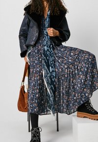 Vestito floreale blu navy con orlo arricciato, abbinato a un giubbotto blu navy cropped con colletto in pelliccia sintetica. Stivali neri con lacci e borsa a tracolla marrone.