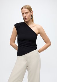 Čierny top s jedným odhaleným ramenom, priliehavého strihu a textúrovanou látkou, doplnený svetlo béžovými širokými nohavicami, na jednoduchom pozadí.